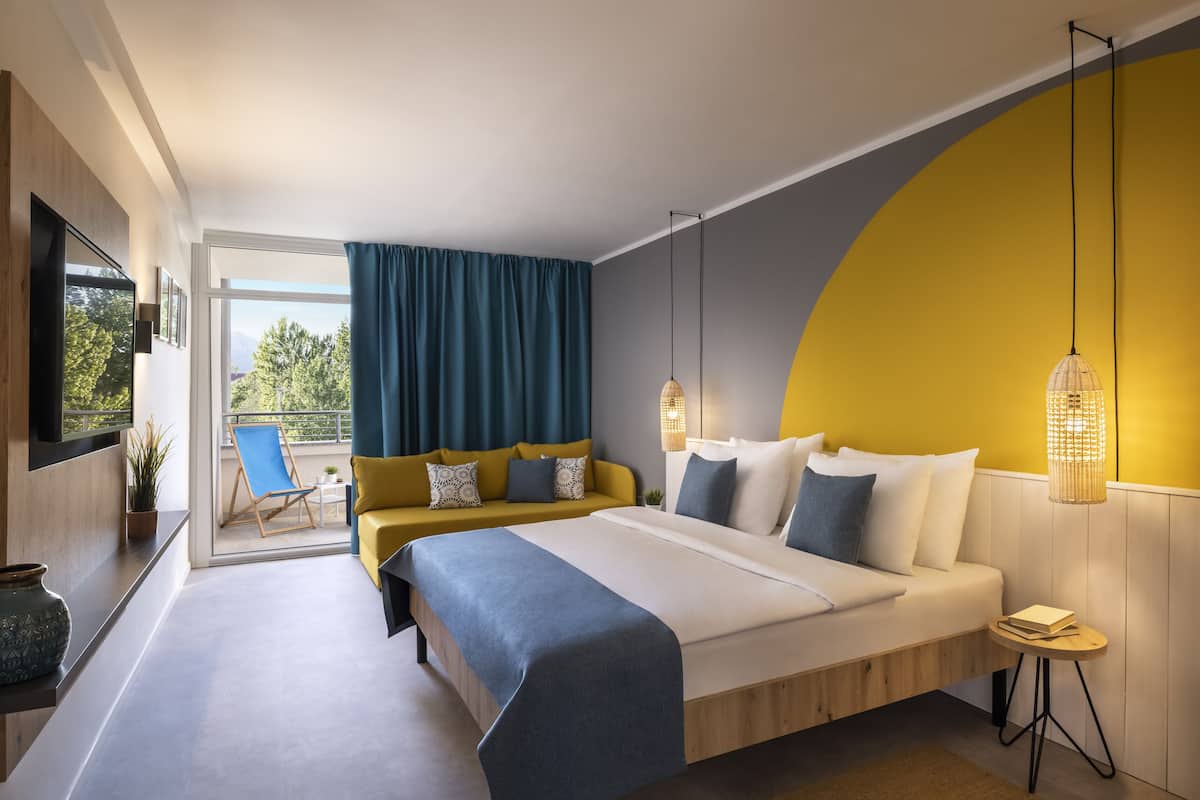 A modern hotel room with a large bed, a balcony with a blue chair, and a yellow accent wall.