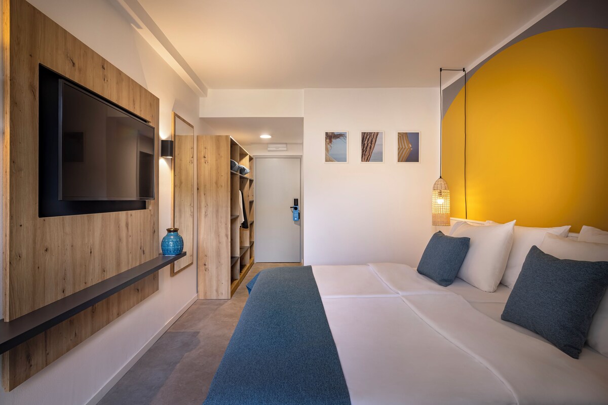 A modern hotel room with a large bed, a flat-screen TV mounted on a wooden panel, a wardrobe, and a small seating area with a lamp.