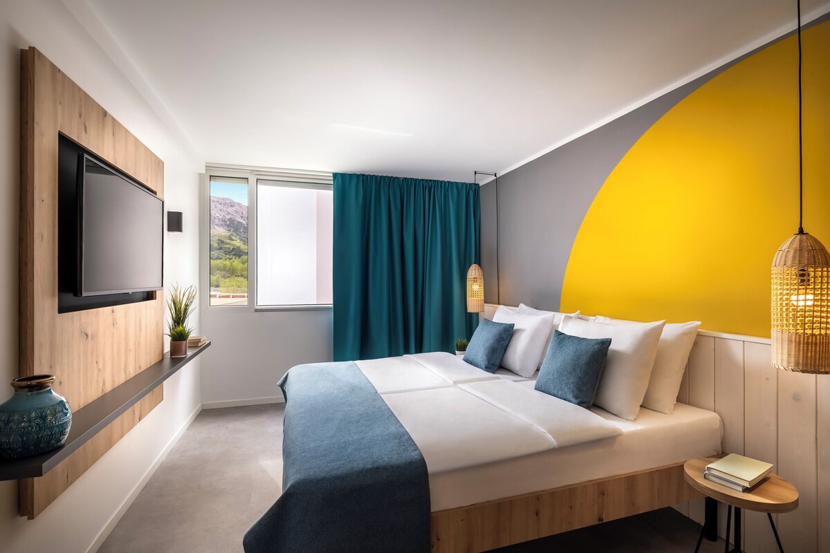 A modern bedroom with a large bed, a TV mounted on the wall, a window with curtains, and a yellow accent wall.