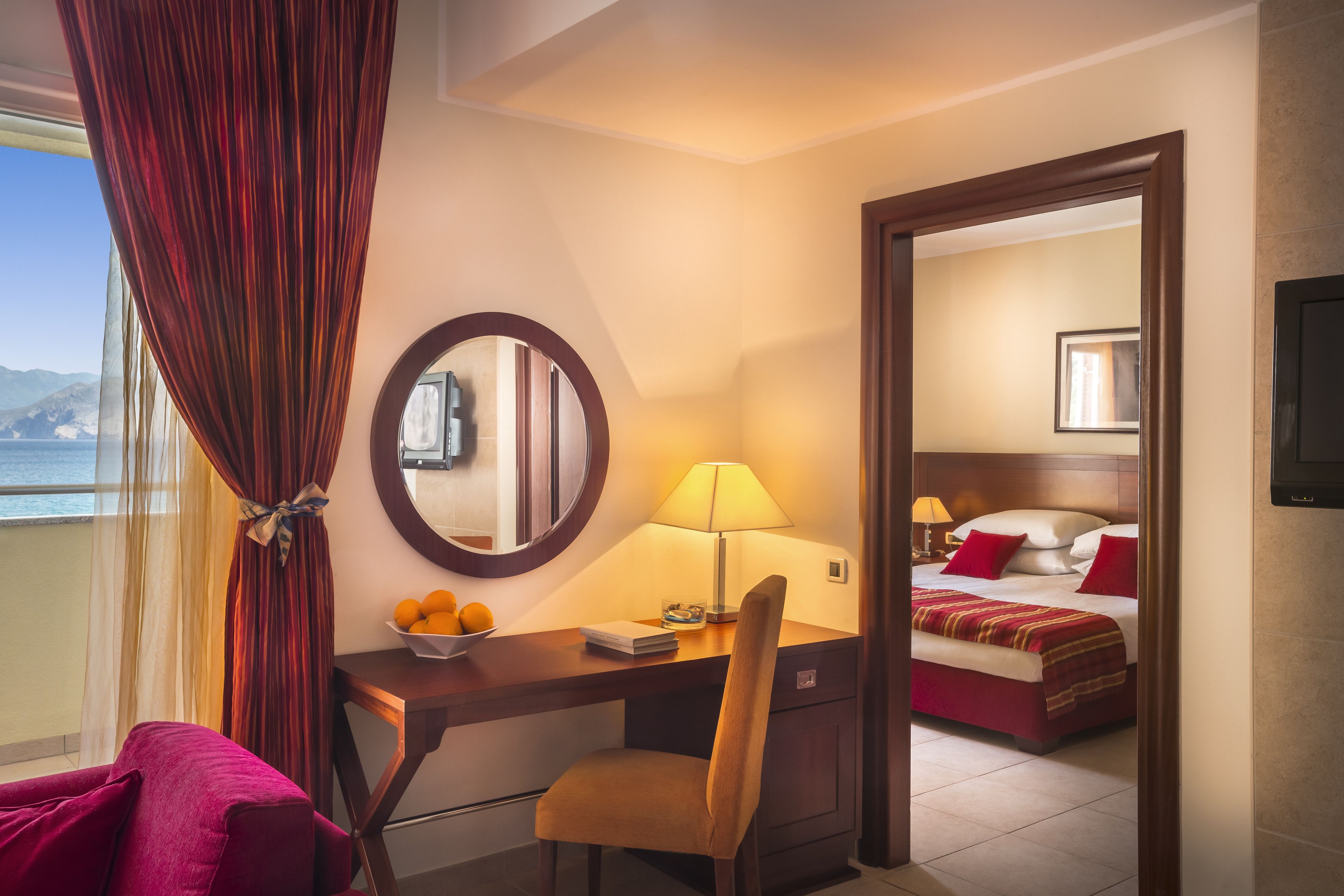 Una habitación de hotel con escritorio, silla y cama con ropa de cama roja. Hay una lámpara en el escritorio y un cuenco de naranjas a un lado.