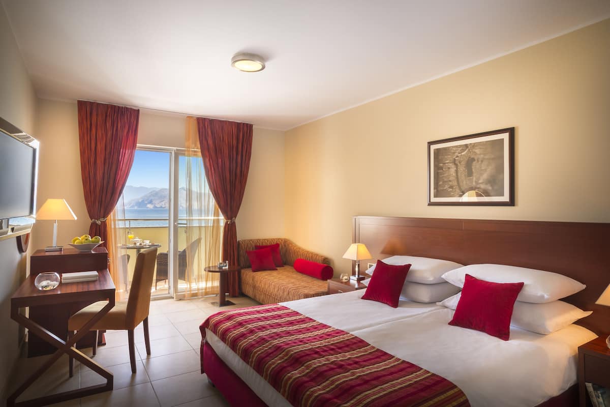 Habitación de hotel con cama, escritorio, silla, televisor y balcón con cortinas.
