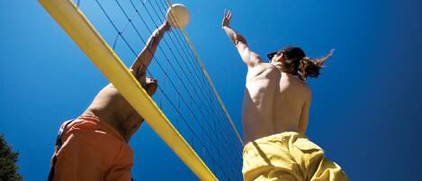 Dos personas jugando voleibol, una vestida de naranja y la otra de amarillo, contra un cielo azul despejado.