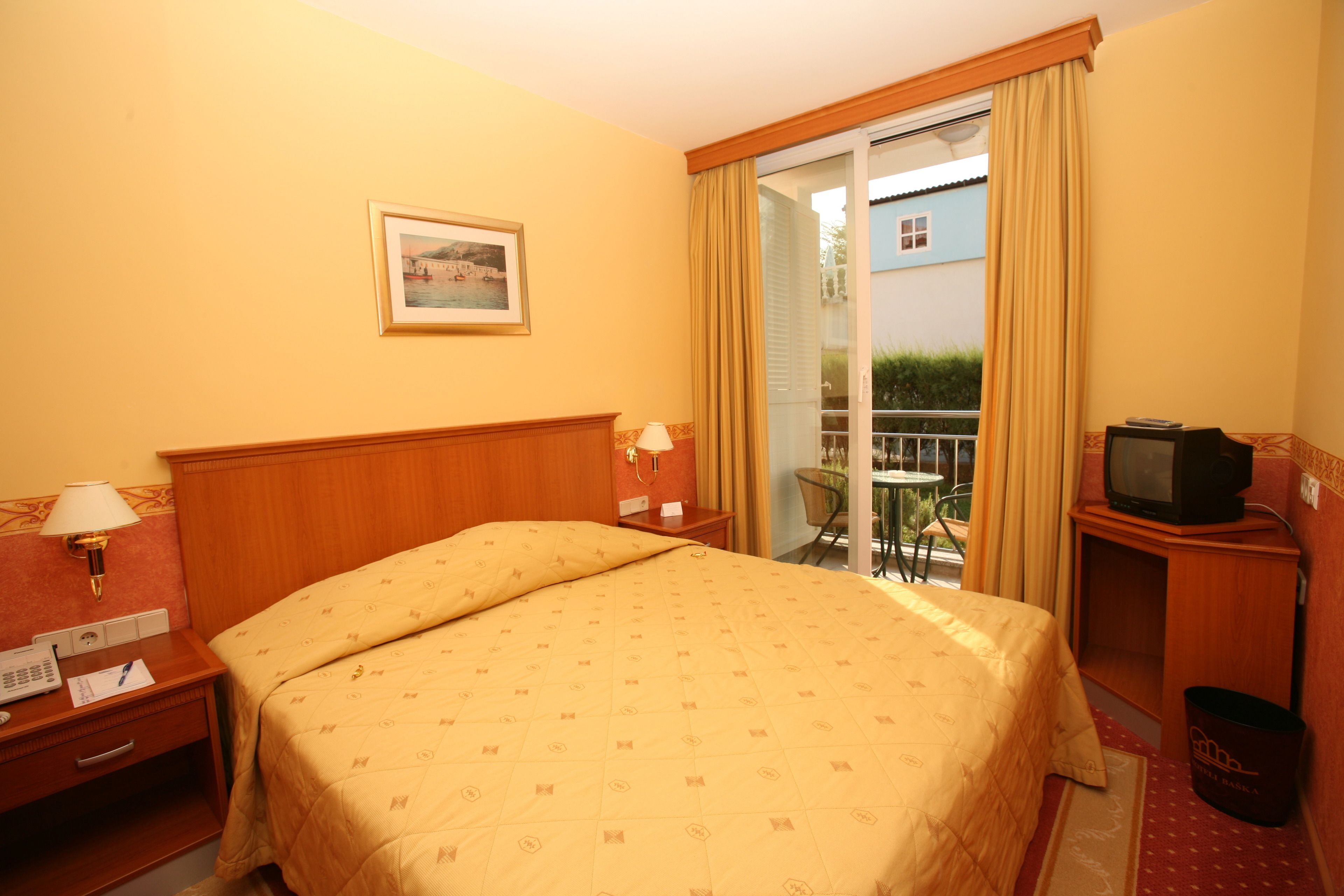 Ein Hotelzimmer mit Bett, Schreibtisch mit Lampe, Fernseher und Blick auf einen Balkon mit Gartenmöbeln.