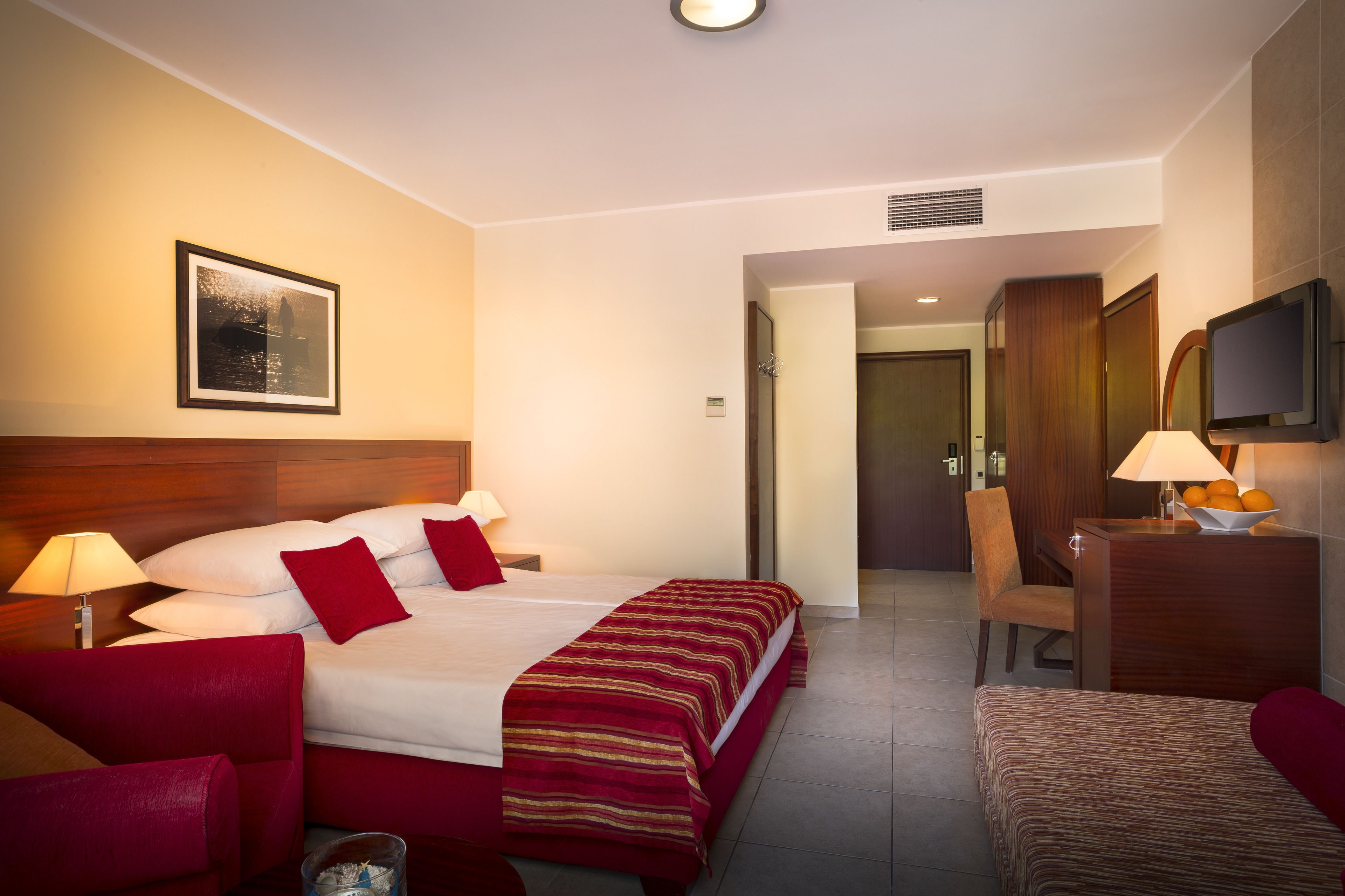A hotel room with a bed, two bedside tables with lamps, a chair, a desk with a lamp, a television, and a framed picture on the wall.