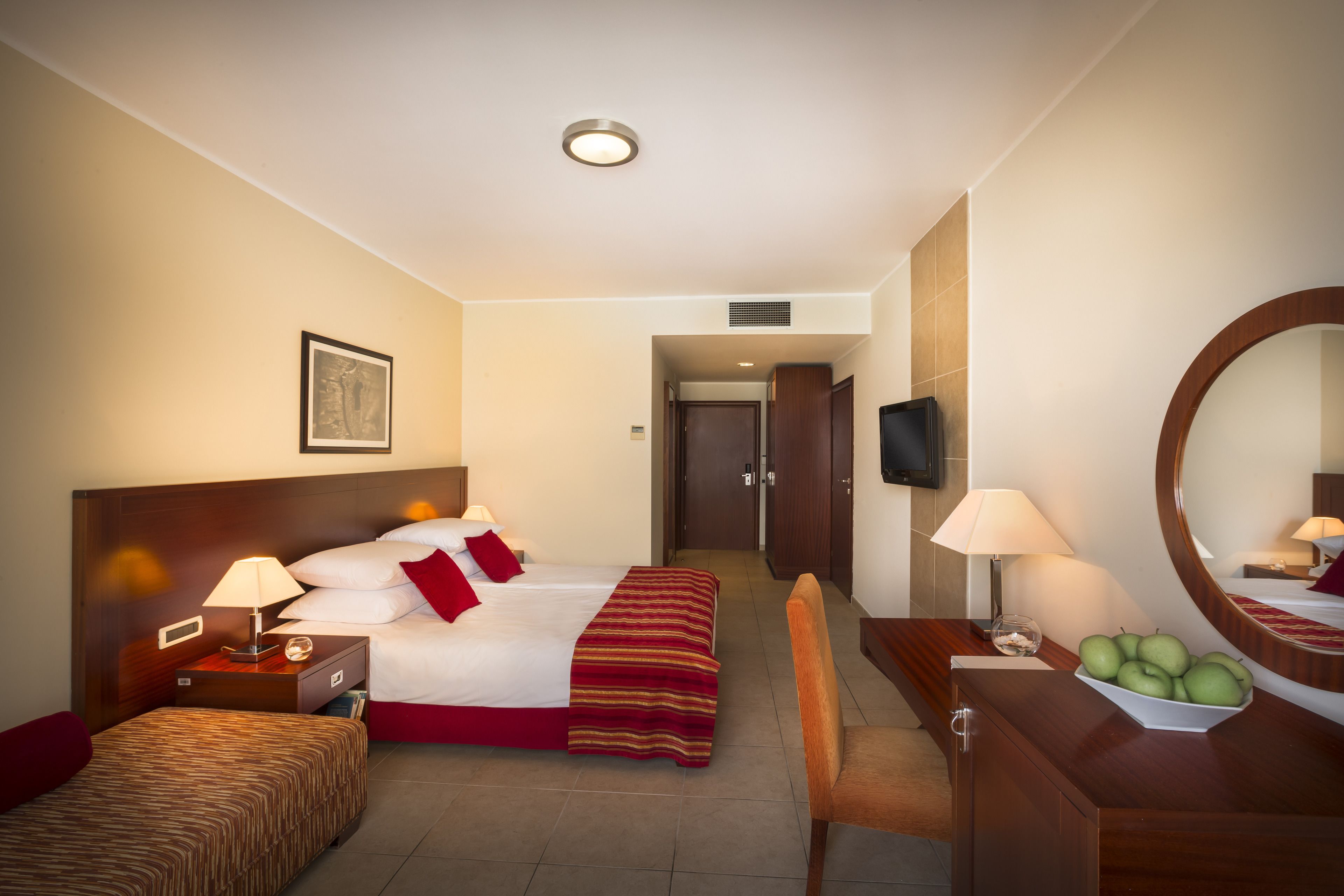 Habitación de hotel con cama, mesita de noche, lámpara, silla, cómoda, espejo y un frutero.