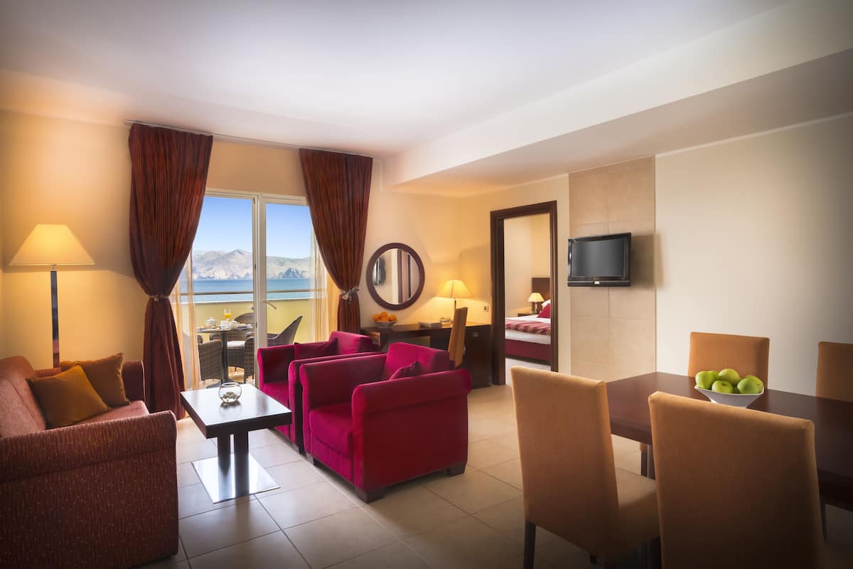 Habitación de hotel con sofá, sillones, una mesa de comedor con sillas, televisor y vistas a las montañas.