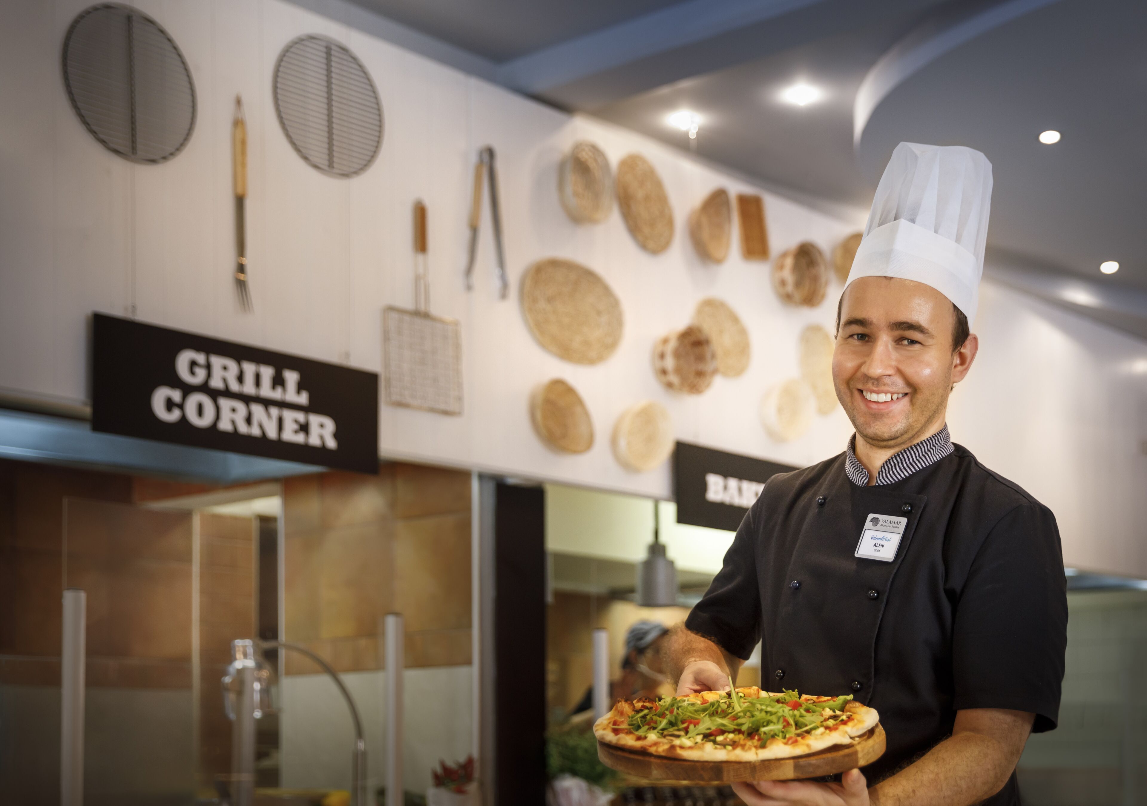 Un chef sostiene una pizza en una cocina con un letrero que dice "GRILL CORNER".