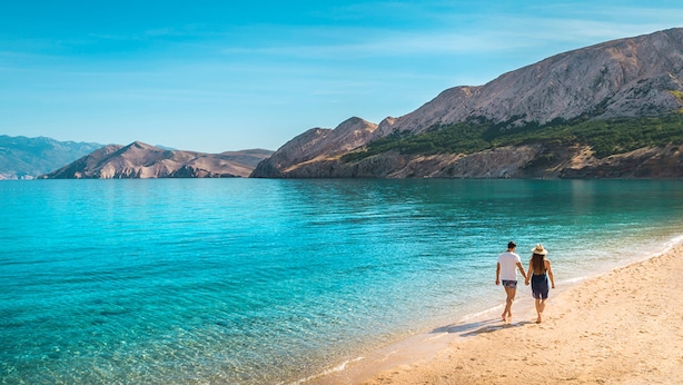 A beach with clear turquoise water and rocky mountains in the background.