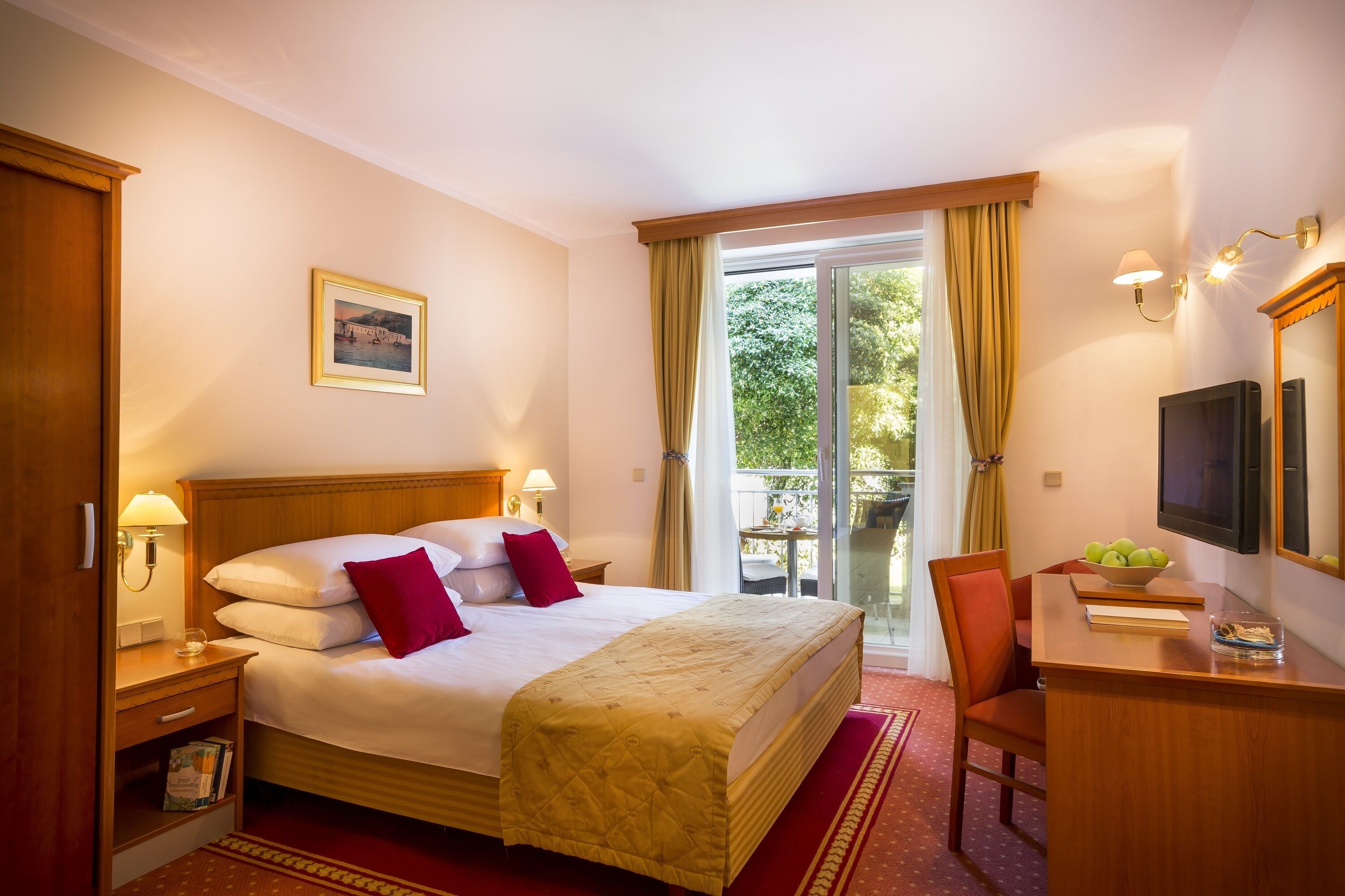 Une chambre d’hôtel avec un grand lit, un bureau avec une télévision, une chaise, une petite table avec un saladier de fruits, une fenêtre avec des rideaux et un tableau au mur.