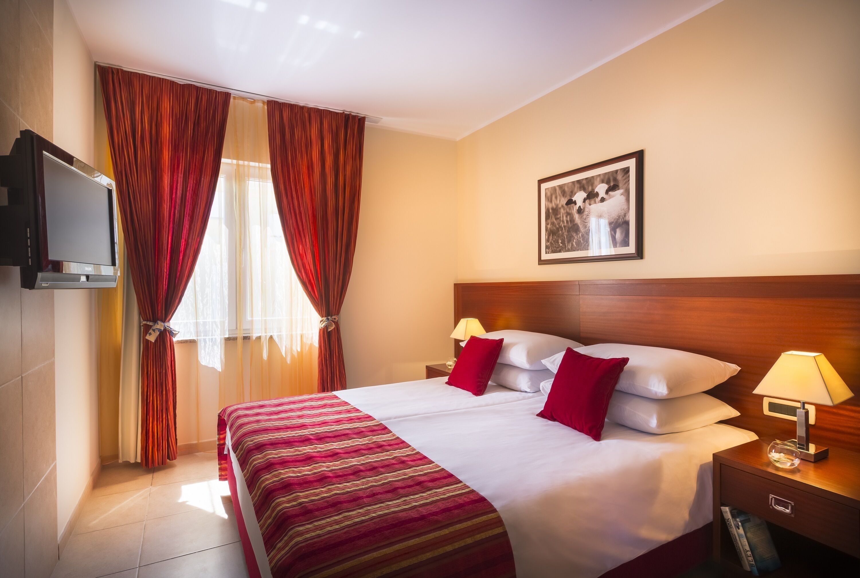 Una camera d'albergo con un letto grande, due cuscini, una lampada da comodino, un televisore montato a parete e un quadro incorniciato.