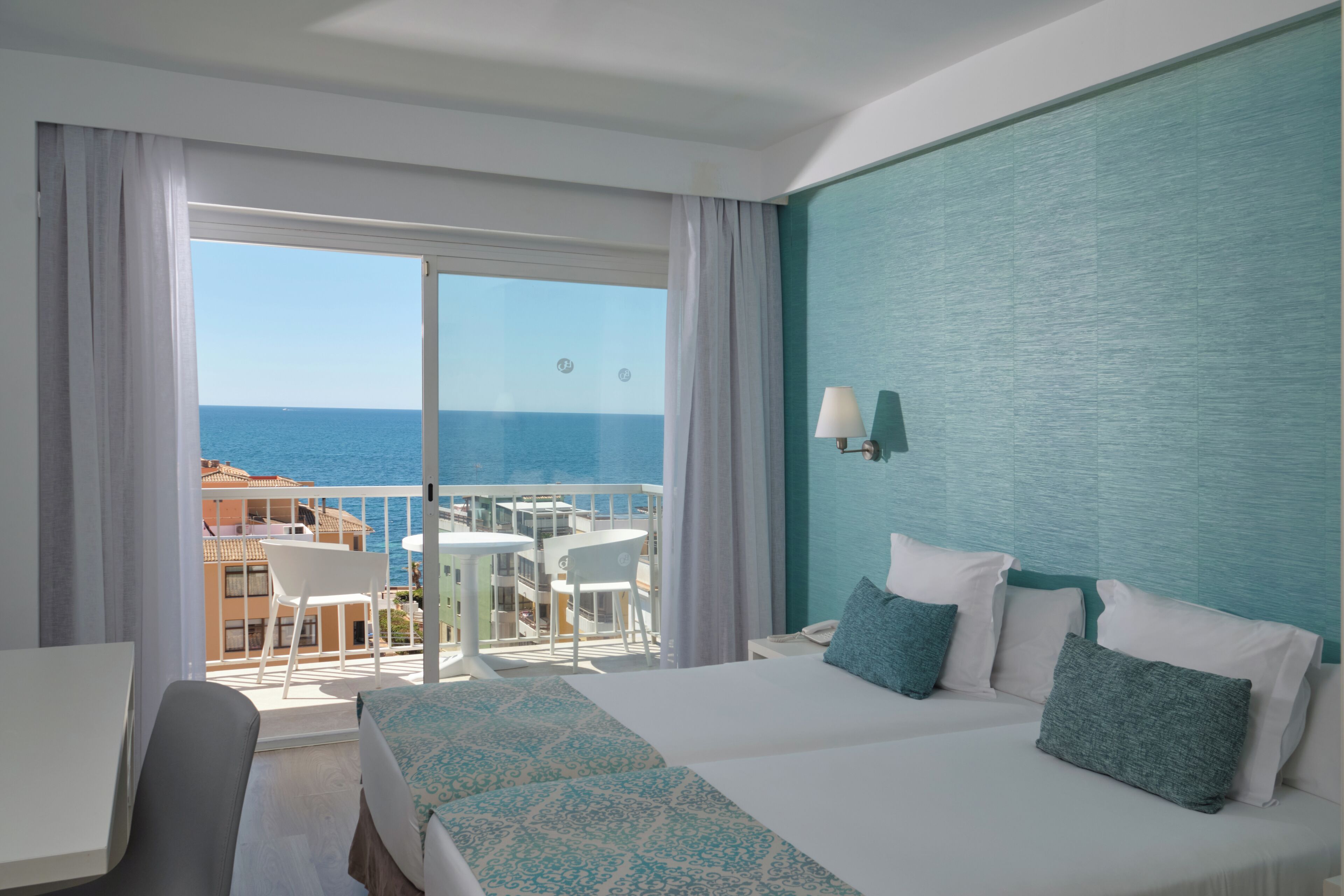 A hotel room with a balcony, a bed, a desk, and a view of the sea.