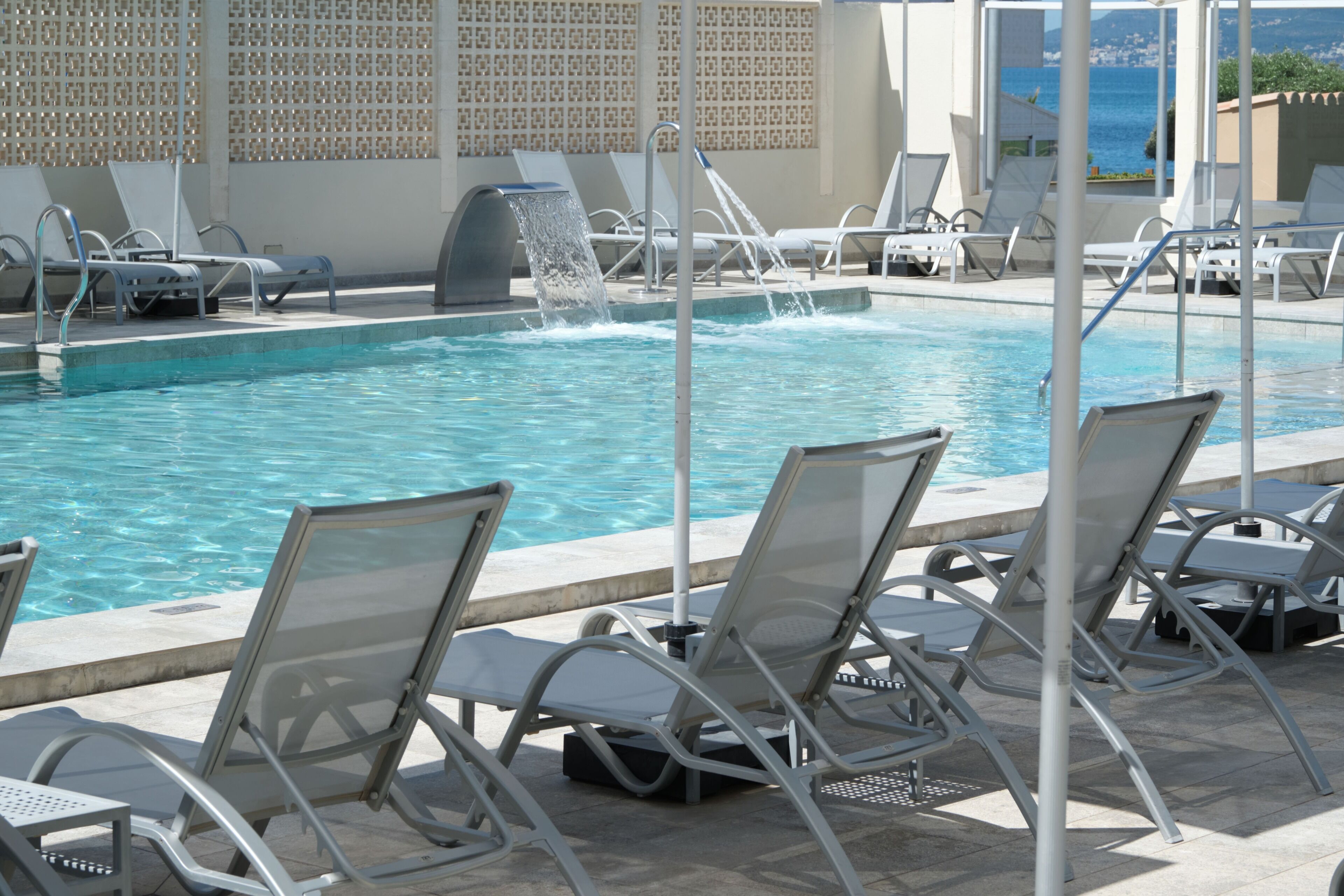 A pool with clear water, surrounded by metal lounge chairs and a wall with a geometric pattern.