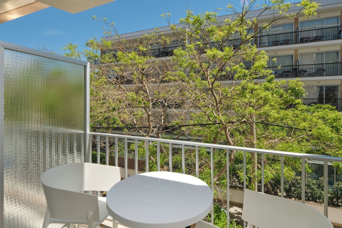 A balcony with white chairs and a table, overlooking a building with balconies and trees.
