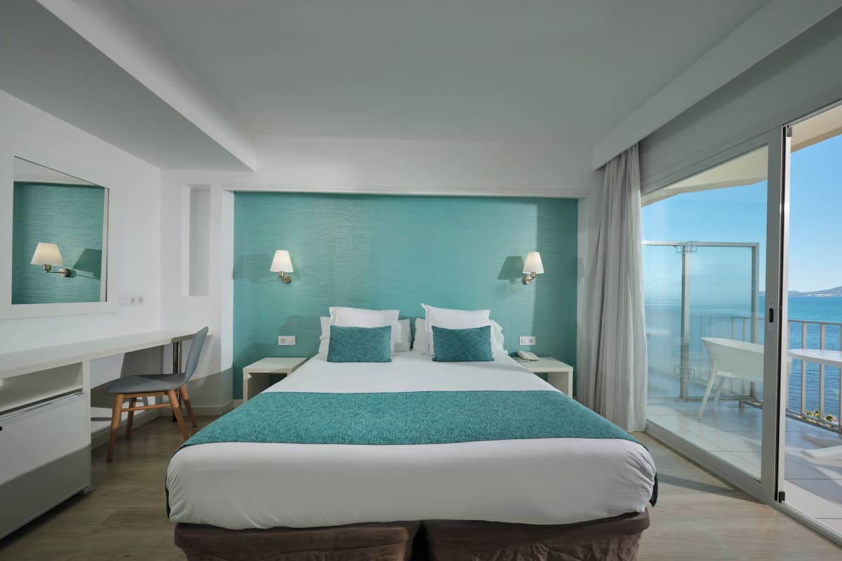 A hotel room with a bed, a desk, a chair, a nightstand, a mirror, and a balcony with a view of the sea.