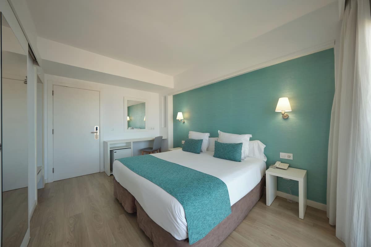 A hotel room with a large bed, a desk, a chair, a mirror, a nightstand, and a lamp.