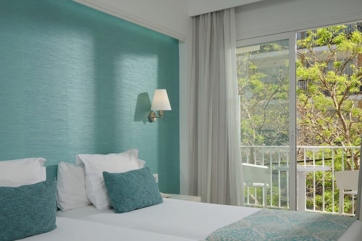 A hotel room with a bed, a window with a view of greenery, and a wall-mounted lamp.