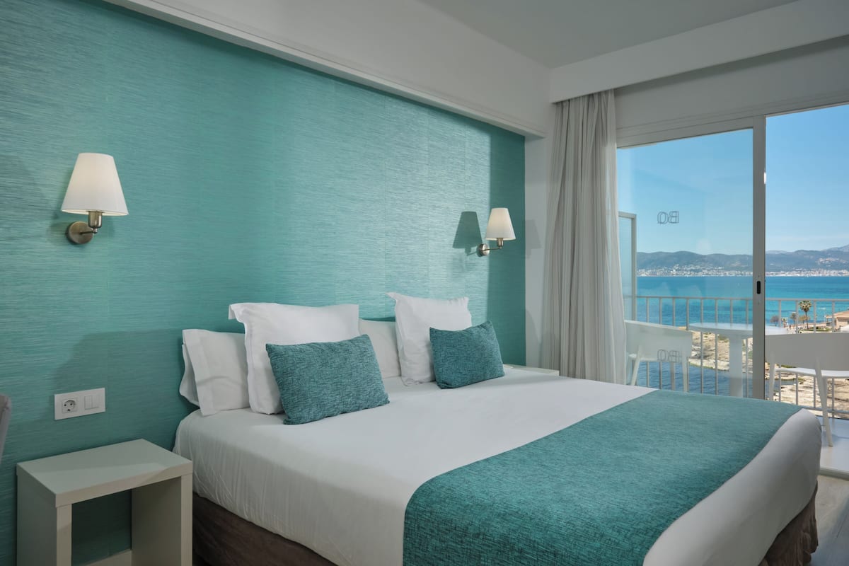 A hotel room with a bed, two bedside tables, a view of the sea, and a balcony with a table and chairs.