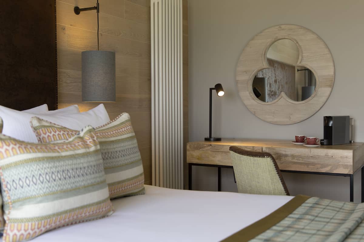 A hotel room with a bed, pillows, a desk, a chair, a lamp, a coffee maker, and a mirror.