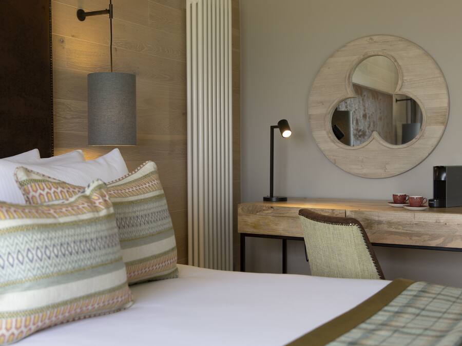 A hotel room with a bed, pillows, a desk, a chair, a lamp, a coffee maker, and a mirror.