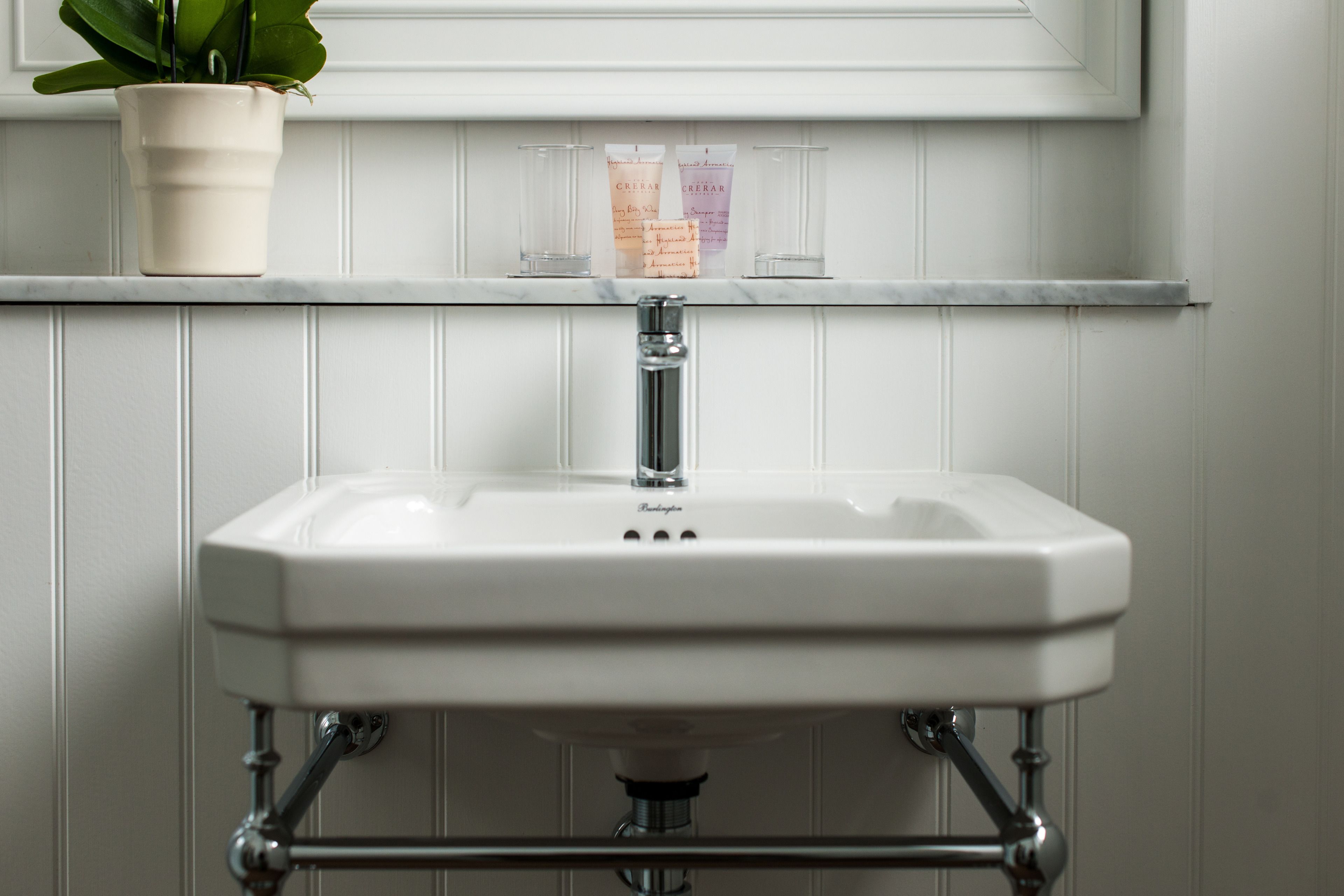 A white bathroom sink with a chrome faucet, a potted plant, and two empty glass bottles on a white shelf above.