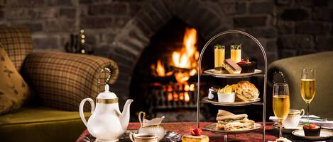 A cozy room with a brick fireplace, a three-tiered tea service, and a variety of snacks and drinks on a table.