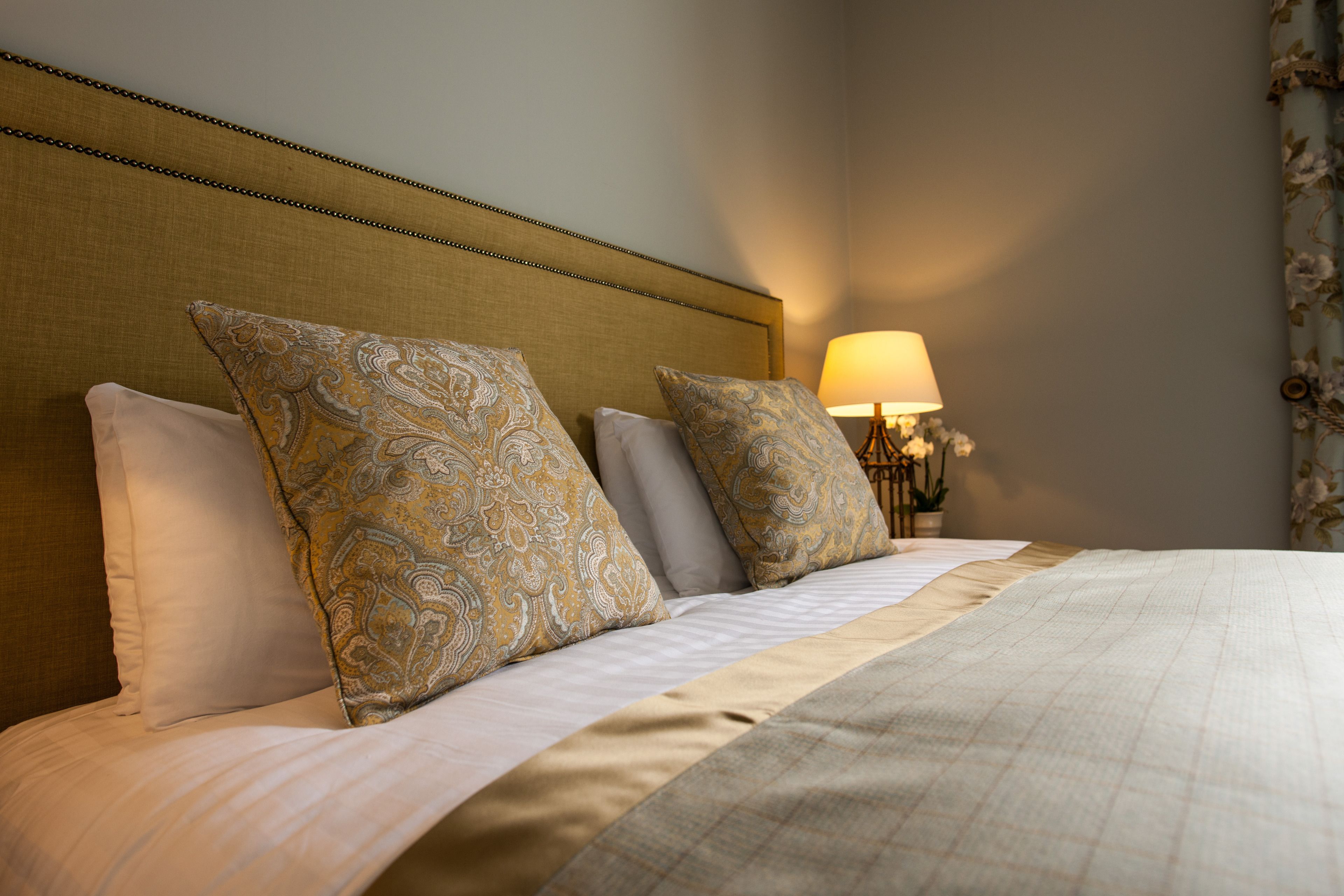 A neatly made bed with patterned pillows and a headboard, a bedside lamp, and a floral curtain.