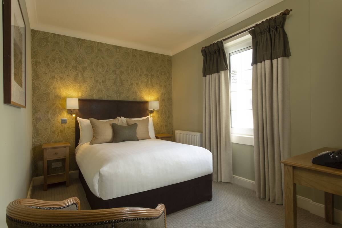 A hotel room with a large bed, bedside lamps, a wooden nightstand, a wicker chair, a wooden table, and a window with curtains.