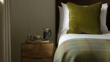Una cama bien tendida con un almohadón verde, una mesita de noche de madera con una cafetera y una taza al lado.