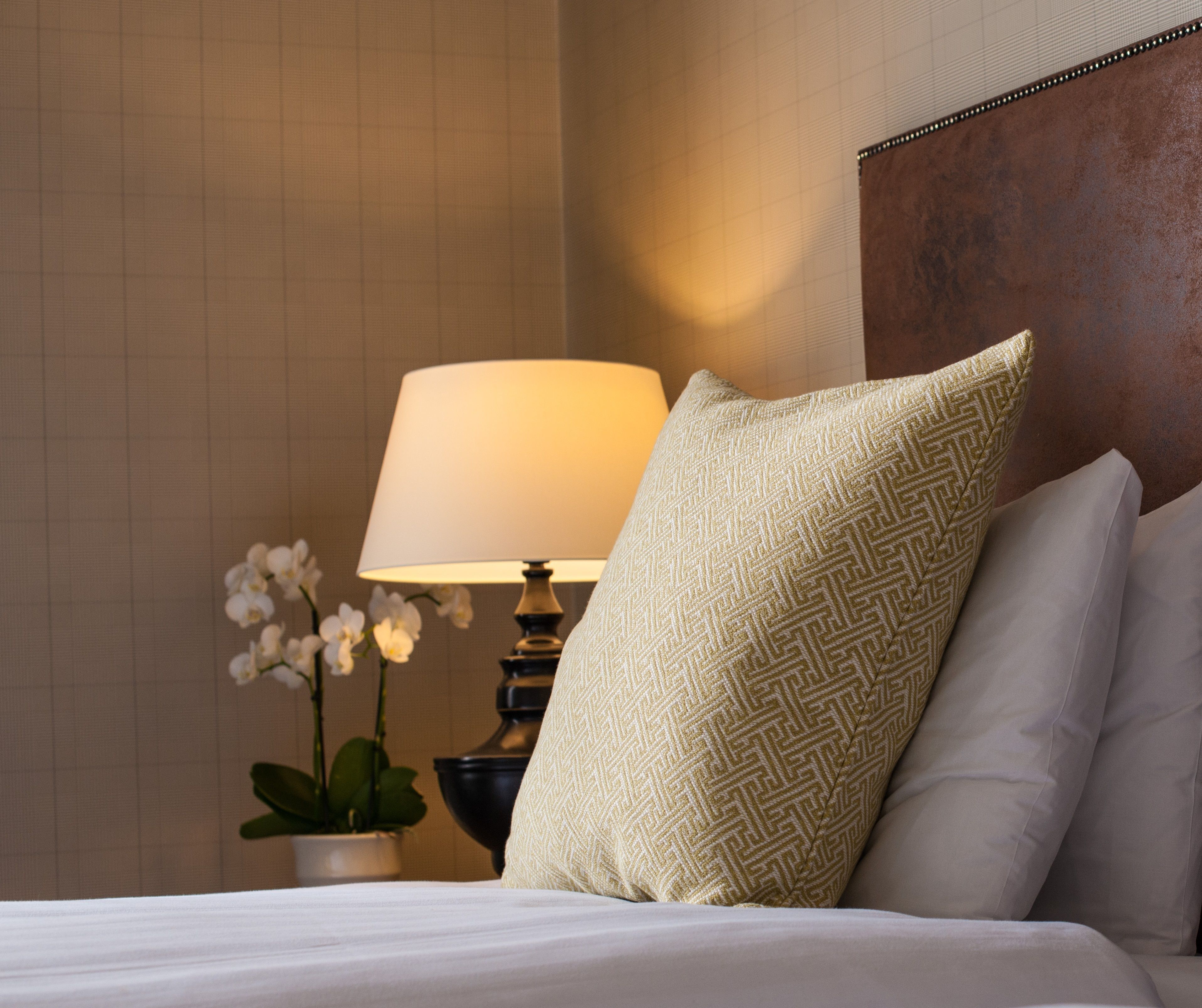 Una cama bien hecha con un cojín estampado, una lámpara de noche y una orquídea en maceta.