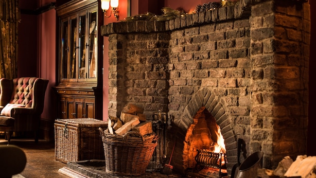 A cozy room with a brick fireplace, a cushioned chair, and a wicker basket filled with logs. - Balmoral Arms (Ballater)