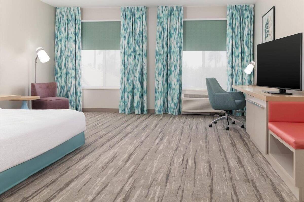 Een hotelkamer met een bed, een bureau met een televisie, twee stoelen, een bureaulamp en een raam met gordijnen.
