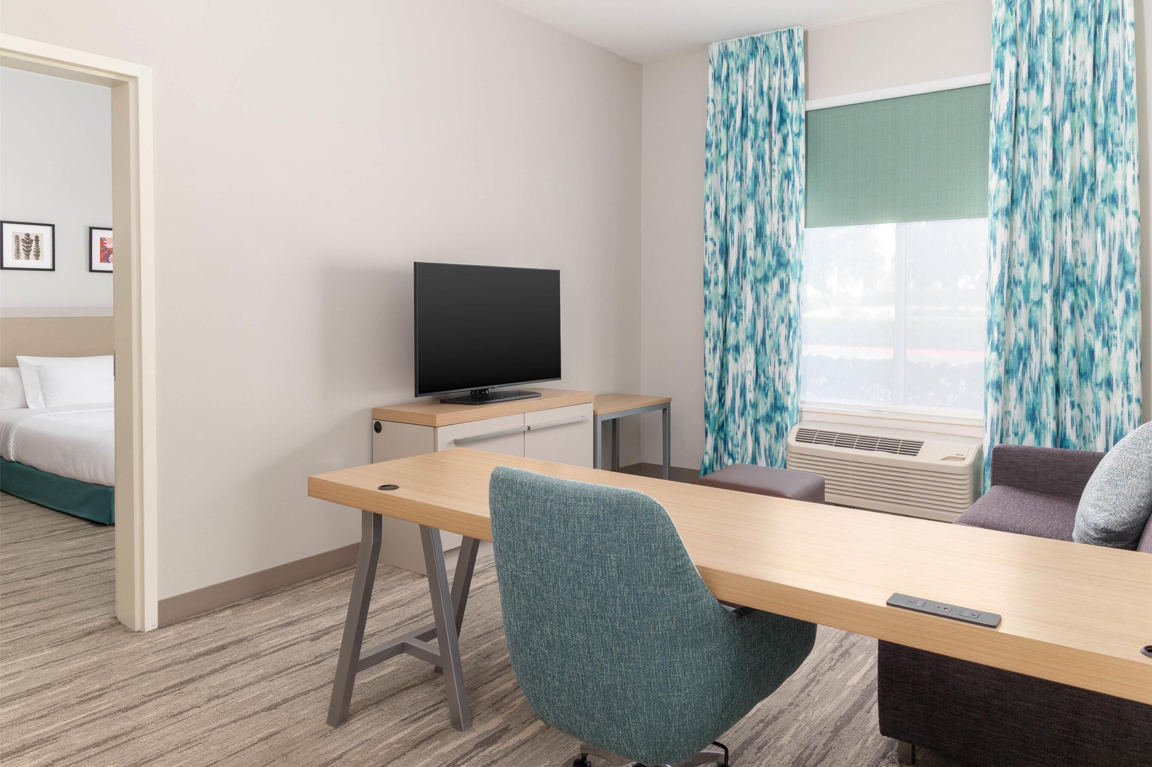 A hotel room with a bed, a desk with a TV, a sofa, and a window with curtains.