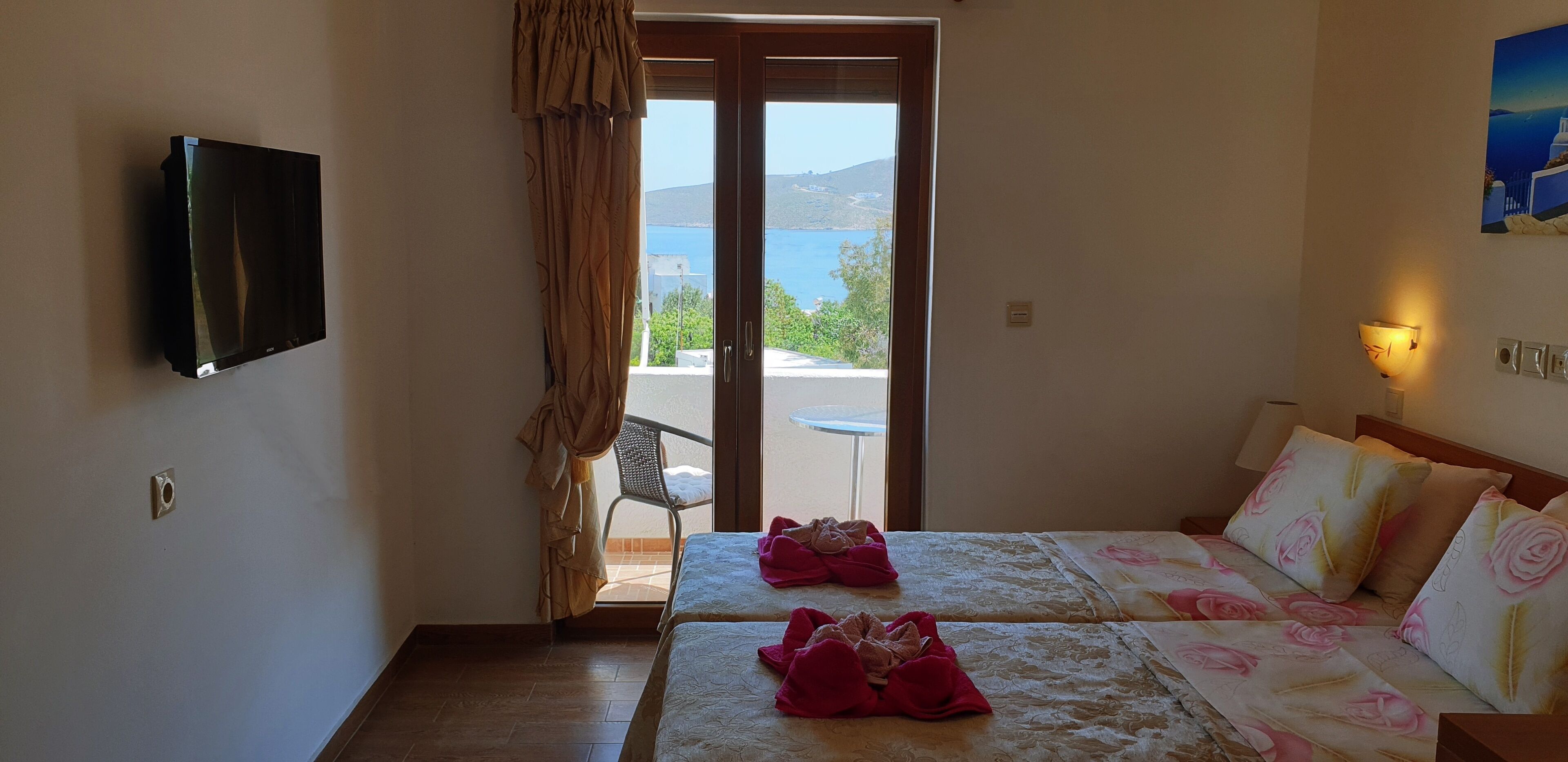 Habitación de hotel con televisión montada en la pared, balcón con vistas al mar y dos camas con ropa de cama estampada.