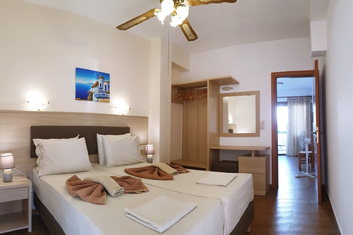 A hotel room with a bed, bedside lamps, a ceiling fan, a painting, a wardrobe, a mirror, and a view of the outside.