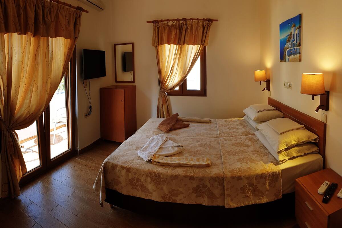 Habitación de hotel con cama, cortinas, televisor, un cuadro y mesita de noche con teléfono y control remoto.