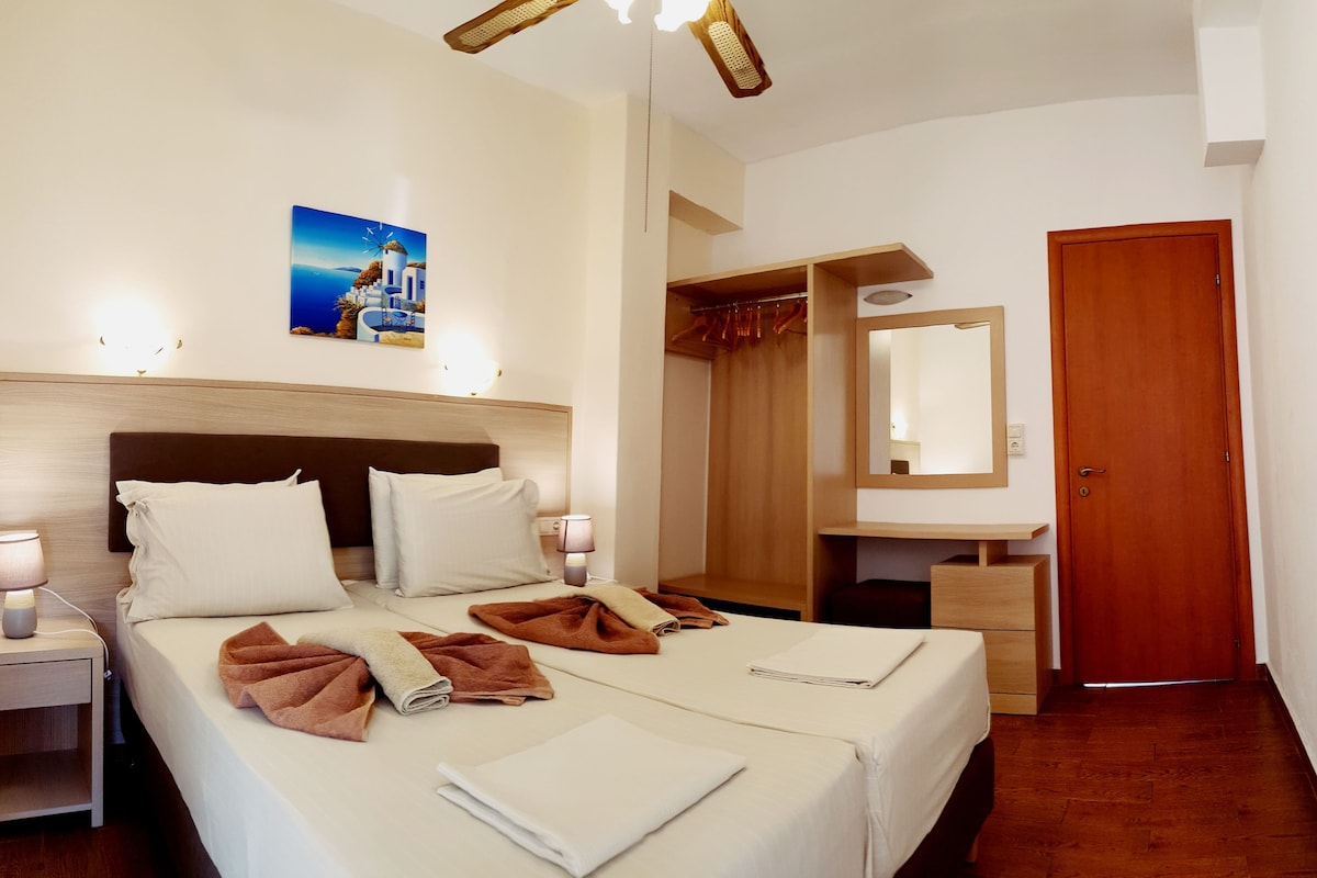 A hotel room with a bed, bedside lamps, a nightstand, a wooden headboard, a wardrobe, a mirror, and a painting of a coastal scene.
