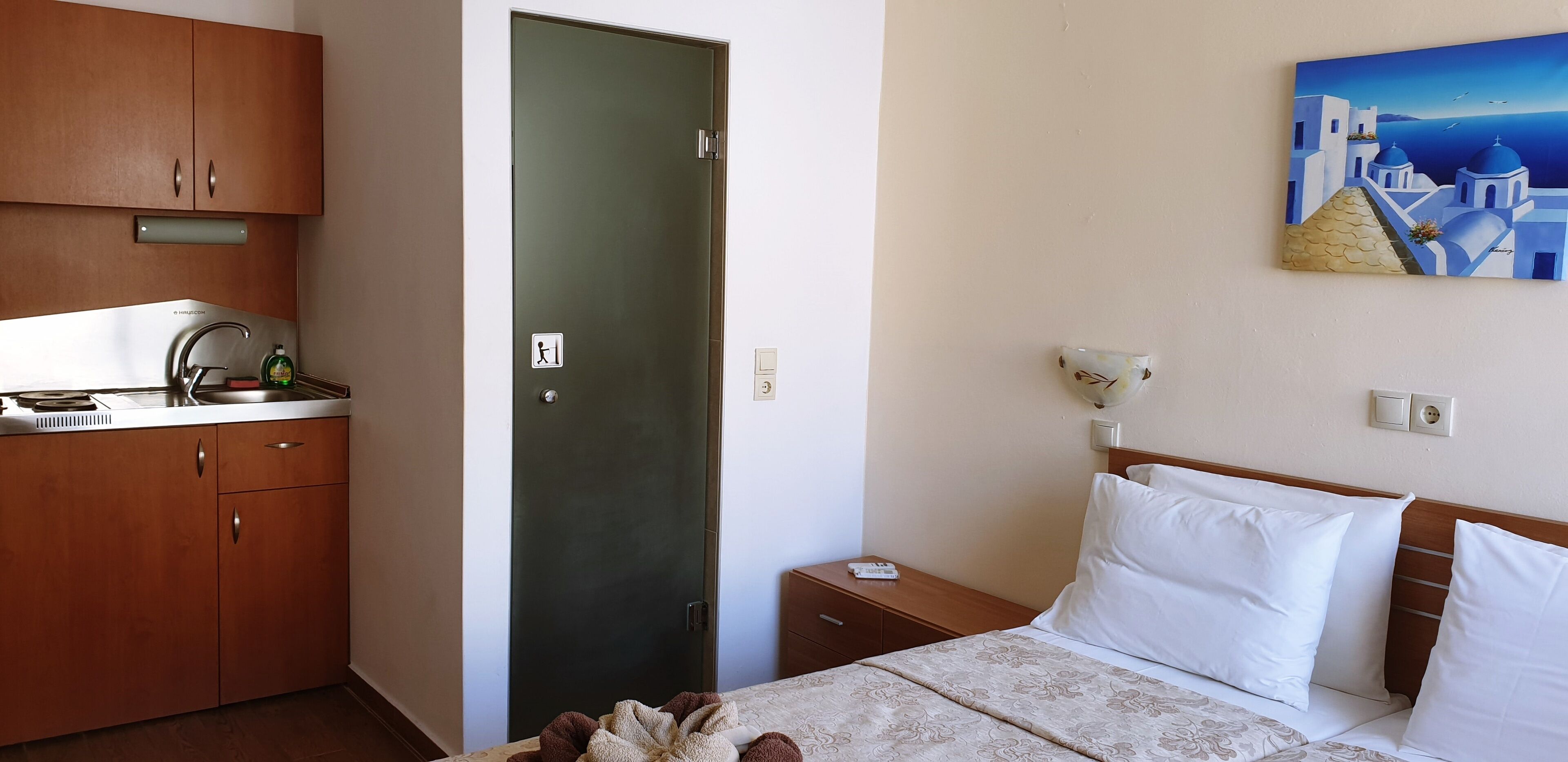 A hotel room with a bed, a nightstand, a painting, and a kitchenette with a sink and cabinets.