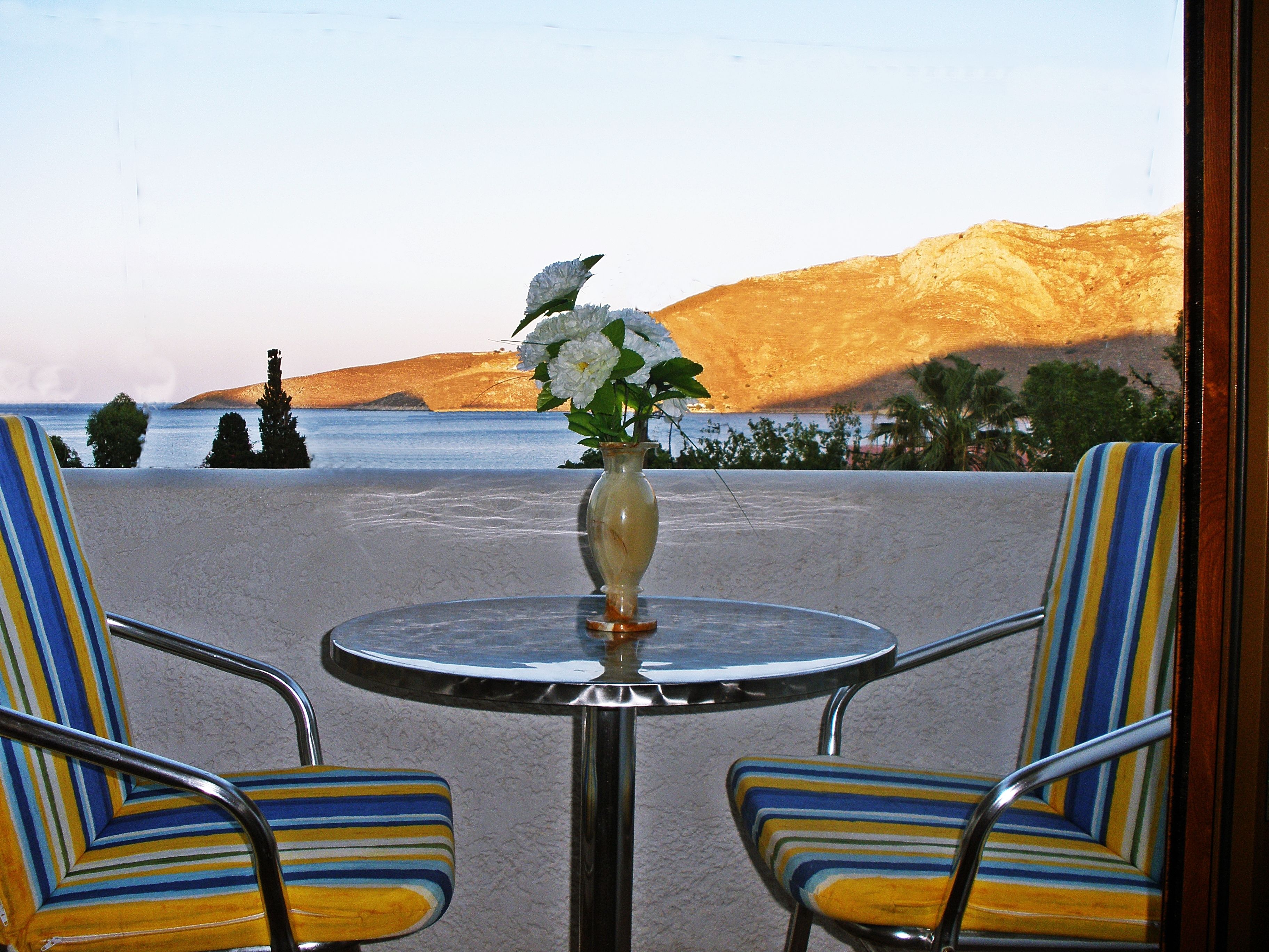 A beachside seating area with a table and chairs, a vase with flowers, and a view of the sea and hills.