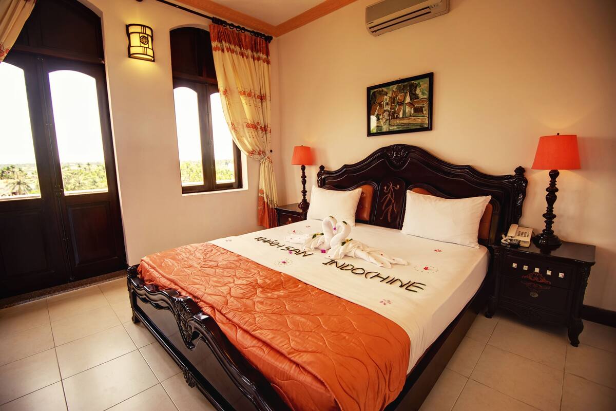 A hotel room with a bed, two bedside lamps, a nightstand, a painting on the wall, and a window with curtains.