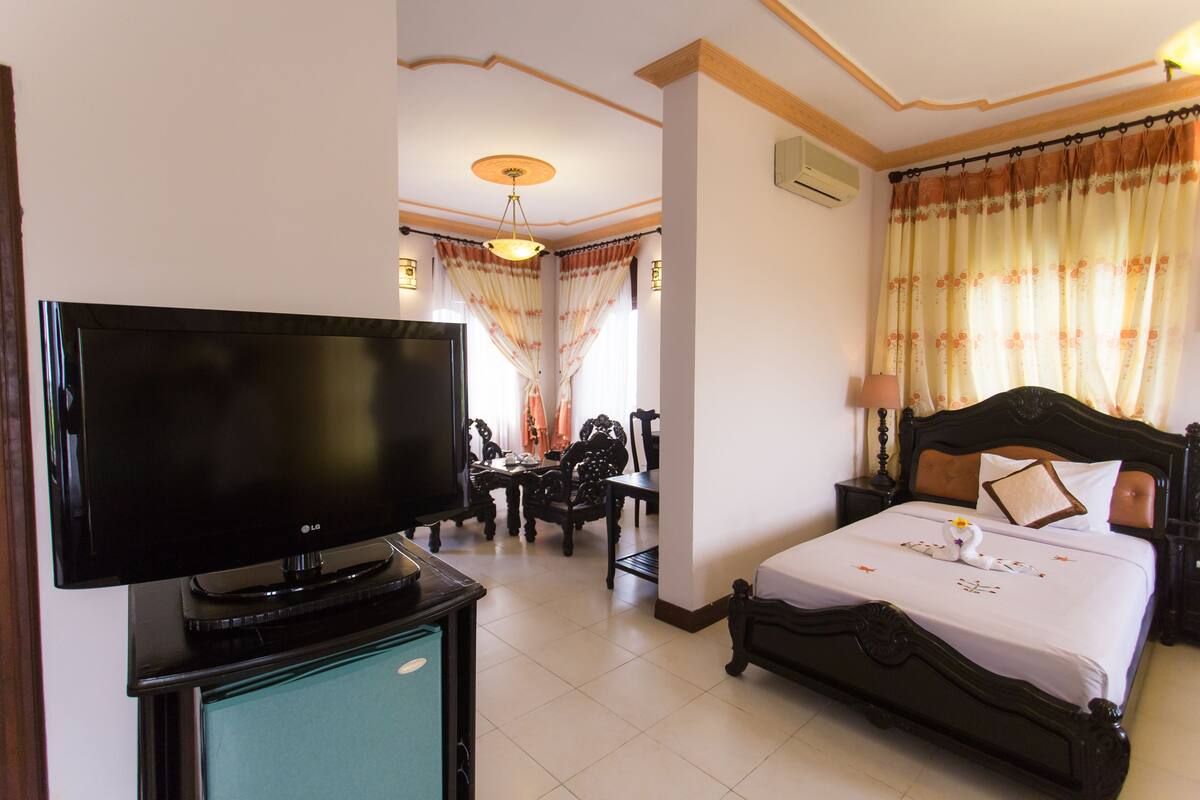 A hotel room with a bed, a television on a stand, a dining table with chairs, and a chandelier.