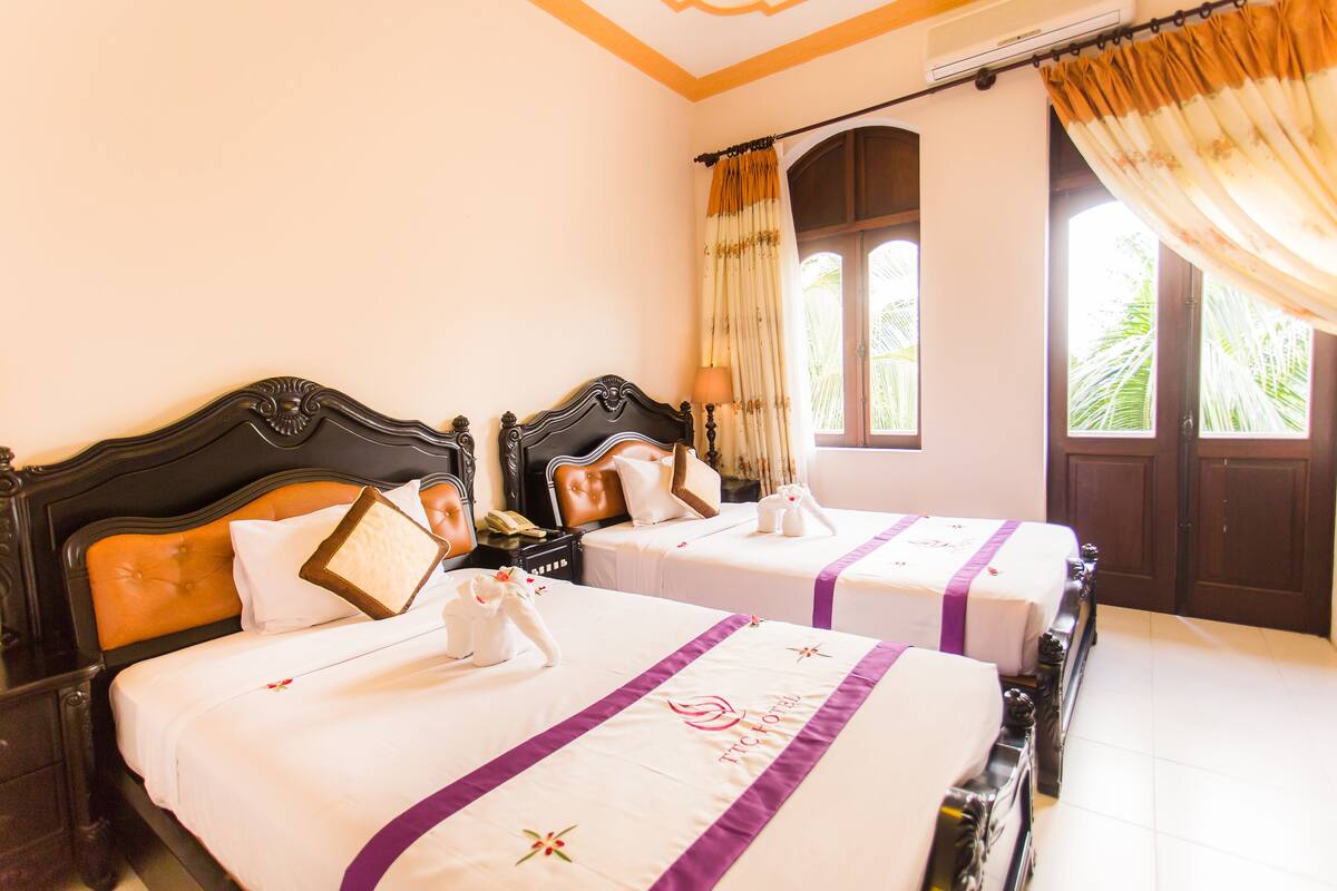 A hotel room with two beds, each with a headboard, a nightstand, a lamp, and a window with curtains.