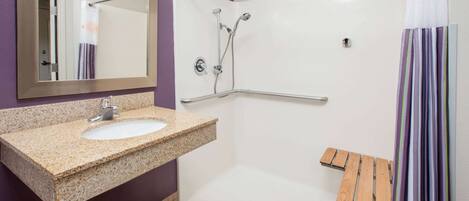 A bathroom with a granite sink, a mirror, a shower with a glass door, and a wooden bench.