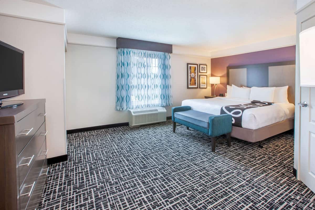 A hotel room with a bed, a desk with a flat-screen TV, a bench, a window with patterned curtains, and a patterned carpet.
