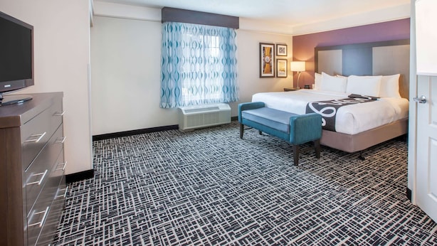 A hotel room with a bed, a desk with a flat-screen TV, a bench, a window with patterned curtains, and a patterned carpet.