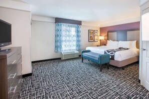 Habitación de hotel con cama, escritorio con televisor de pantalla plana, banco, ventana con cortinas estampadas y alfombra con diseño.