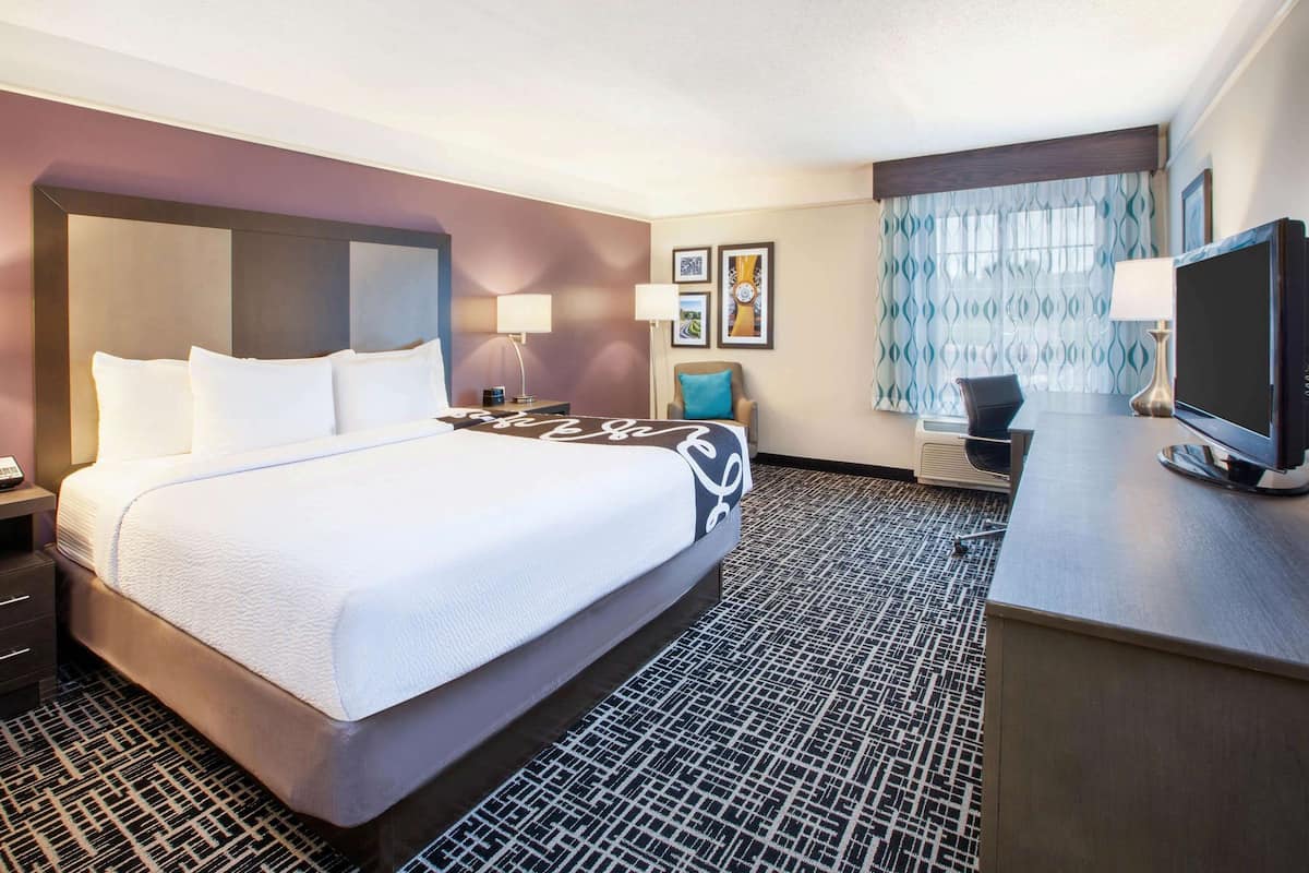 A hotel room with a large bed, a desk with a television, and a window with patterned curtains.