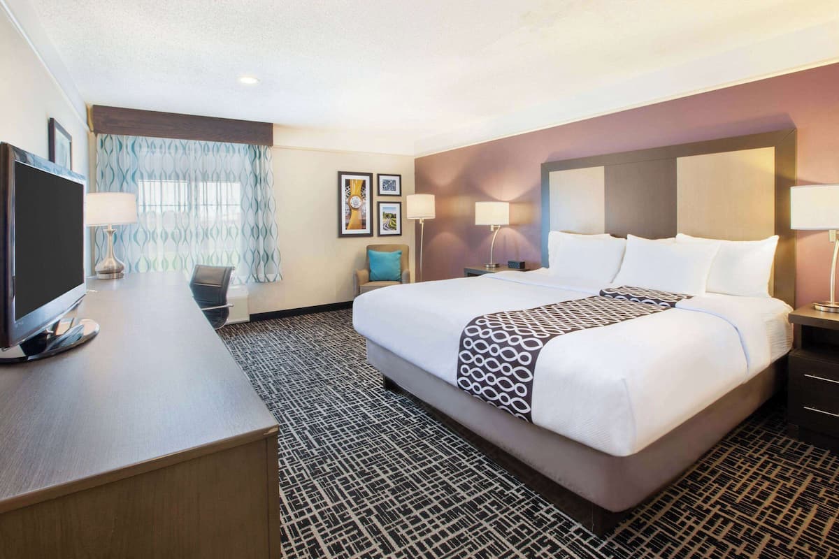 A hotel room with a large bed, a desk with a flat-screen TV, a chair, and a window with patterned curtains.