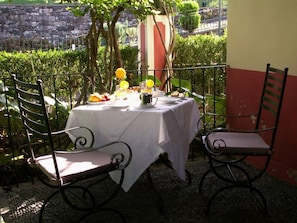 Un espace de restauration en plein air avec une table dressée pour deux personnes, entouré de chaises en métal, d’un mur en pierre et d’une végétation en arrière-plan.