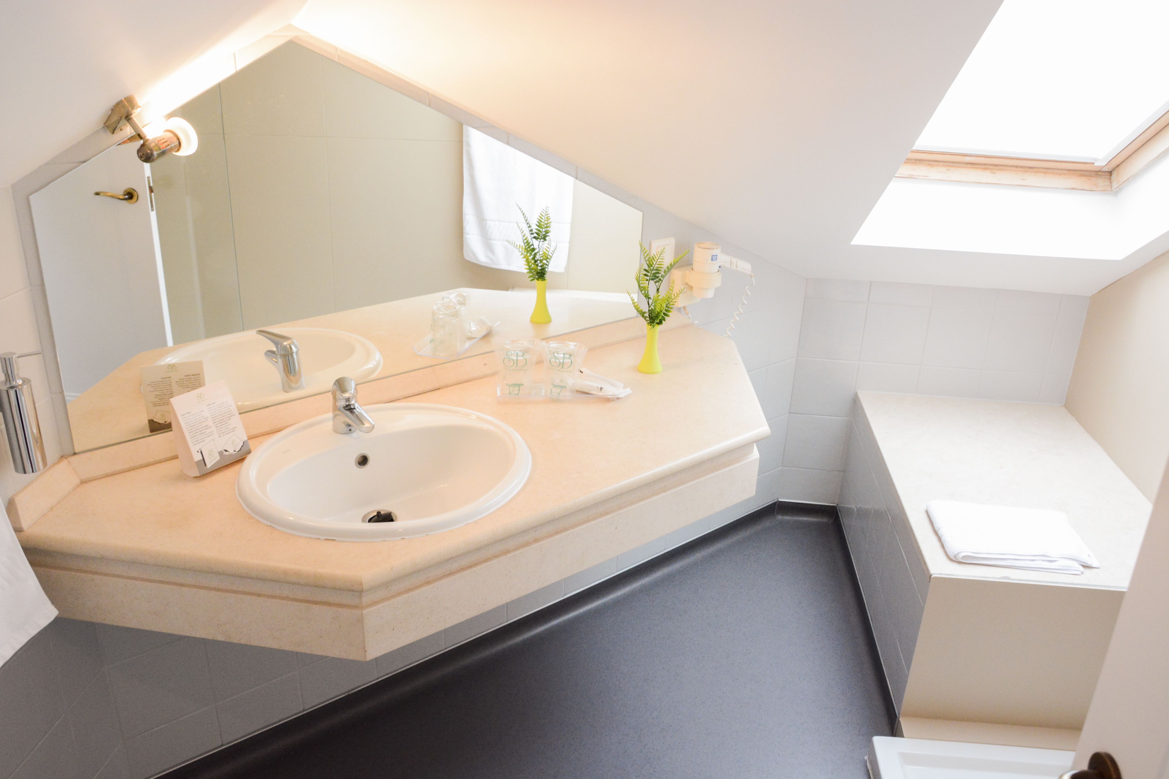 A modern bathroom with a large mirror, two sinks, a bathtub, and a skylight.