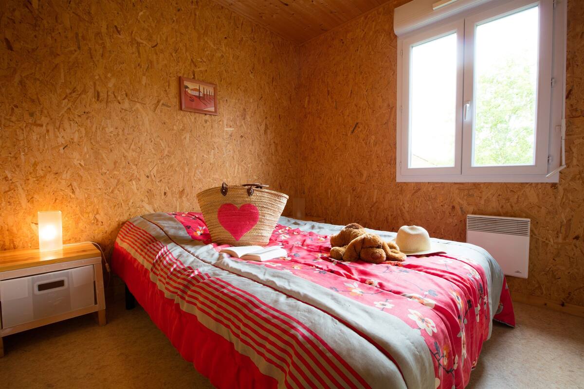 Une chambre avec un mur en bois, un lit recouvert d’une courtepointe à motifs floraux, un ours en peluche, un panier et une table de chevet avec une lampe.